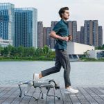 Eine Person joggt auf einem Holzsteg am Wasser, während der vierbeinige Unitree Go2 Roboter seitlich neben ihr mitläuft; im Hintergrund stehen moderne Hochhäuser und Grünflächen.